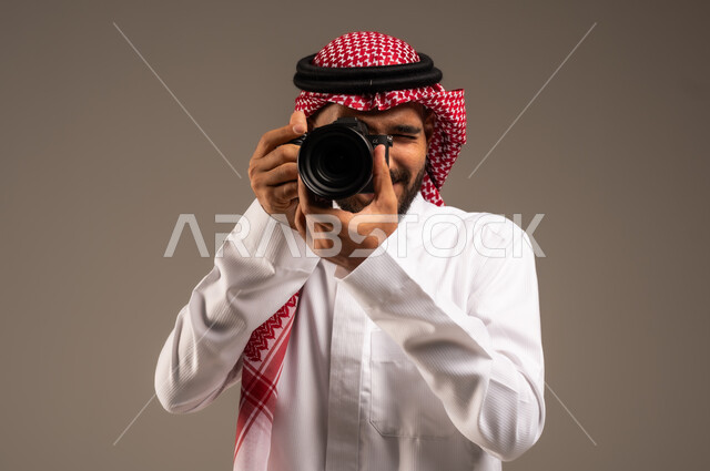 استخدام كاميرات رقمية عالية الدقة، إتقان واحتراف مهنة التصوير، بورتريه مقرب لرجل عربي خليجي سعودي يرتدي الشماغ والثوب التقليدي يقوم بالتقاط صور تذكارية عبر كاميرا فوتوغرافية، وظائف ومهن سعودية، خلفية رمادية