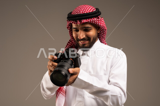 إيماءات الإعجاب والسعادة باحتراف المهنة، مصور فوتوغرافي يقوم بمراجعة لقطاته، بورتريه مقرب لشاب عربي خليجي سعودي مبتسم يرتدي الثوب التقليدي والشماغ يحمل بيده كاميرا التصوير عالية الدقة، معدات وتقنيات متطورة، خلفية رمادية