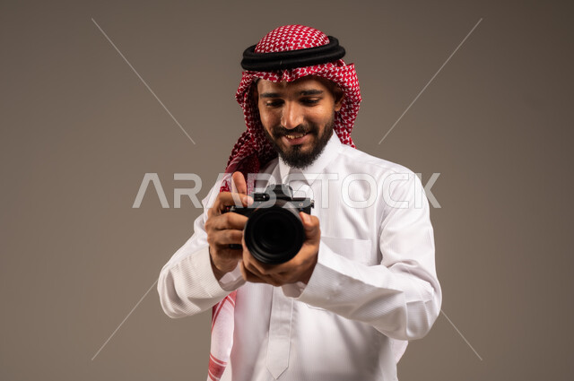إيماءات الإعجاب والسعادة باحتراف المهنة، مصور فوتوغرافي يقوم بمراجعة لقطاته، بورتريه مقرب لشاب عربي خليجي سعودي مبتسم يرتدي الثوب التقليدي والشماغ يحمل بيده كاميرا التصوير عالية الدقة، معدات وتقنيات متطورة، خلفية رمادية