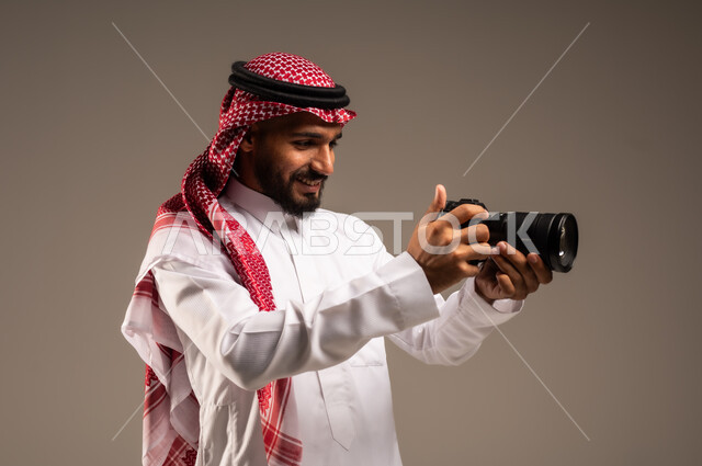 إيماءات الإعجاب والسعادة باحتراف المهنة، مصور فوتوغرافي يقوم بمراجعة لقطاته، بورتريه مقرب لشاب عربي خليجي سعودي مبتسم يرتدي الثوب التقليدي والشماغ يحمل بيده كاميرا التصوير عالية الدقة، معدات وتقنيات متطورة، خلفية رمادية