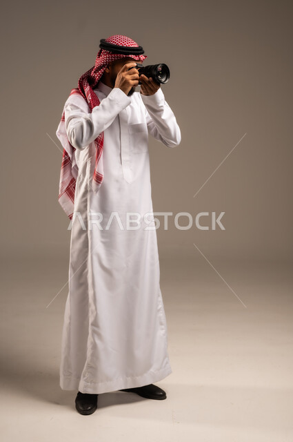 إتقان واحتراف مهنة التصوير، بورتريه لرجل عربي خليجي سعودي يرتدي الشماغ والثوب التقليدي يقوم بالتقاط صور تذكارية عبر كاميرا فوتوغرافية، استخدام كاميرات رقمية عالية الدقة، وظائف ومهن سعودية، صورة الجسم بالطول الكامل، خلفية رمادية