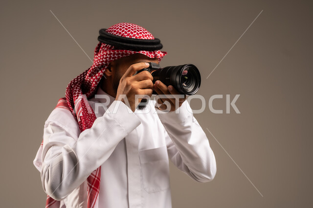 إتقان واحتراف مهنة التصوير، بورتريه مقرب لرجل عربي خليجي سعودي يرتدي الشماغ والثوب التقليدي يقوم بالتقاط صور تذكارية عبر كاميرا فوتوغرافية، استخدام كاميرات رقمية عالية الدقة، وظائف ومهن سعودية، خلفية رمادية