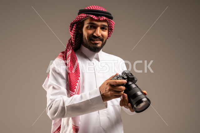 إيماءات الإعجاب والسعادة باحتراف المهنة، معدات وتقنيات متطورة، بورتريه مقرب لشاب عربي خليجي سعودي مبتسم يرتدي الثوب التقليدي والشماغ يحمل بيده كاميرا للتصوير الفوتوغرافي عالي الدقة وينظر إلى الكاميرا، خلفية رمادية