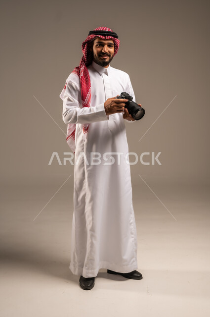 Gestures of admiration and happiness with professionalism, advanced equipment and techniques, portrait of a smiling young Saudi Arabian Gulf man wearing traditional thobe and shemagh holding a high-resolution photography camera and looking into the camera, full-length body image, gray background