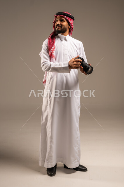 معدات وتقنيات متطورة، بورتريه لشاب عربي خليجي سعودي مبتسم يرتدي الثوب التقليدي والشماغ يحمل بيده كاميرا وينظر إلى شيء ما، التصوير الفوتوغرافي عالي الدقة، إيماءات الإعجاب والسعادة باحتراف المهنة، صورة الجسم بالطول الكامل، خلفية رمادية