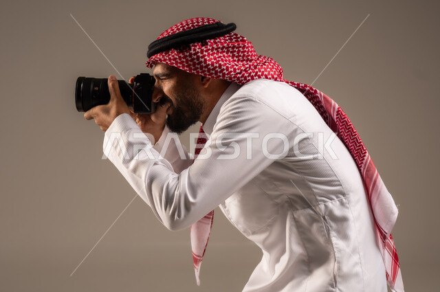 Using high-resolution digital cameras, mastery and professionalism in the photography profession, Saudi jobs and professions, close-up portrait from the side of a Saudi Arabian Gulf man wearing a traditional shemagh and thobe taking souvenir photos with a photographic camera, gray background