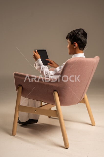 استخدام الأجهزة التقنية الحديثة، بورتريه من الخلف لطفل عربي سعودي خليجي يرتدي الثوب التقليدي يجلس على كرسي مريح ويقوم باستخدام التابلت، مفهوم التعليم وإنجاز المهام عن بعد، قضاء وقت ممتع على الإنترنت، اللعب على الجهاز اللوحي، خلفية رمادية