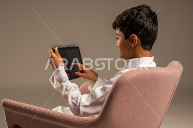 The concept of distance learning and completing tasks, using modern technical devices, close-up portrait from the back of a Saudi Arabian Gulf child wearing traditional dress sitting on a comfortable chair and using a tablet, having fun on the internet, playing on the tablet, gray background