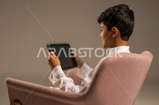  قضاء وقت ممتع على الإنترنت، اللعب على الجهاز اللوحي، بورتريه مقرب من الخلف لطفل عربي سعودي خليجي يرتدي الثوب التقليدي يجلس على كرسي مريح ويقوم باستخدام التابلت، مفهوم التعليم وإنجاز المهام عن بعد، استخدام الأجهزة التقنية الحديثة، خلفية رمادية