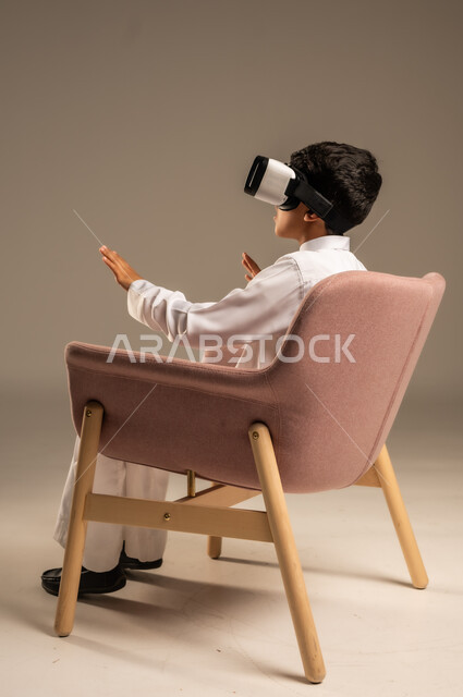 تعابير الاندماج بالعالم الخيالي، بورتريه من الخلف لفتى عربي خليجي سعودي يرتدي الثوب التقليدي يجلس على كرسي مريح ويرتدي نظارة الواقع الافتراضي ثلاثية الابعاد، قضاء أوقات ممتعة ومسلية بالألعاب، خلفية رمادية