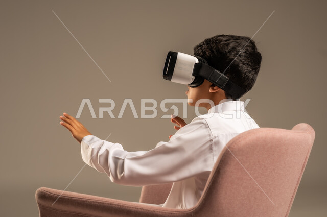 قضاء أوقات ممتعة ومسلية بالألعاب، تعابير الاندماج بالعالم الخيالي، بورتريه مقرب من الخلف لفتى عربي خليجي سعودي يرتدي الثوب التقليدي يجلس على كرسي مريح ويرتدي نظارة الواقع الافتراضي ثلاثية الابعاد، خلفية رمادية