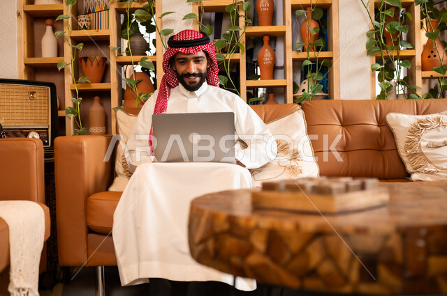  مفهوم إدارة شؤون الأعمال وتنظيمها عبر الاجهزة الحديثة، صورة مقربة لرجل عربي سعودي خليجي يرتدي الثوب التقليدي والشماغ يجلس في  محل لبيع الهدايا التذكارية التقليدية يقوم بالعمل على الحاسوب المحمول، مهن ووظائف مكتبية 