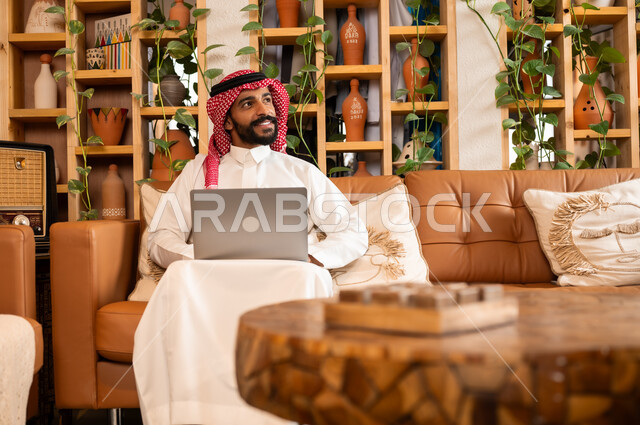  مفهوم إدارة شؤون الأعمال وتنظيمها عبر الاجهزة الحديثة، صورة مقربة لرجل عربي سعودي خليجي يرتدي الثوب التقليدي والشماغ يجلس في  محل لبيع الهدايا التذكارية التقليدية يقوم بالعمل على الحاسوب المحمول، مهن ووظائف مكتبية 