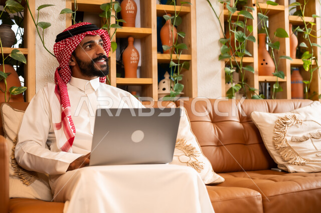  مفهوم إدارة شؤون الأعمال وتنظيمها عبر الاجهزة الحديثة، صورة مقربة لرجل عربي سعودي خليجي يرتدي الثوب التقليدي والشماغ يجلس في  محل لبيع الهدايا التذكارية التقليدية يقوم بالعمل على الحاسوب المحمول، مهن ووظائف مكتبية 