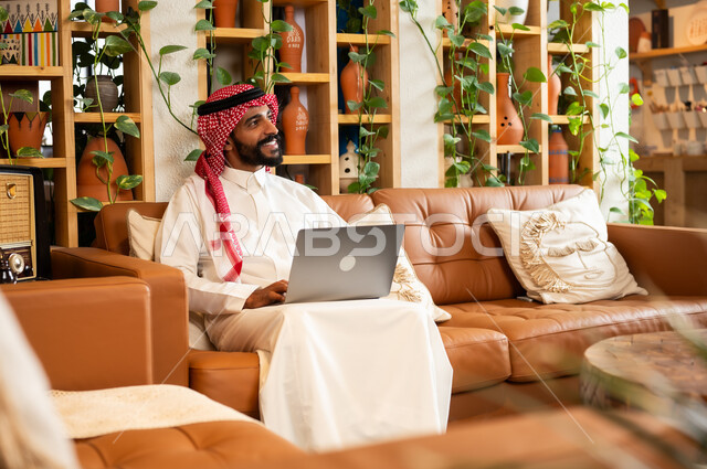  مفهوم إدارة شؤون الأعمال وتنظيمها عبر الاجهزة الحديثة، صورة مقربة لرجل عربي سعودي خليجي يرتدي الثوب التقليدي والشماغ يجلس في  محل لبيع الهدايا التذكارية التقليدية يقوم بالعمل على الحاسوب المحمول، مهن ووظائف مكتبية 