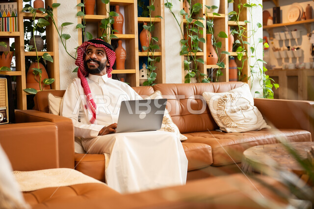  مفهوم إدارة شؤون الأعمال وتنظيمها عبر الاجهزة الحديثة، صورة مقربة لرجل عربي سعودي خليجي يرتدي الثوب التقليدي والشماغ يجلس في  محل لبيع الهدايا التذكارية التقليدية يقوم بالعمل على الحاسوب المحمول، مهن ووظائف مكتبية 