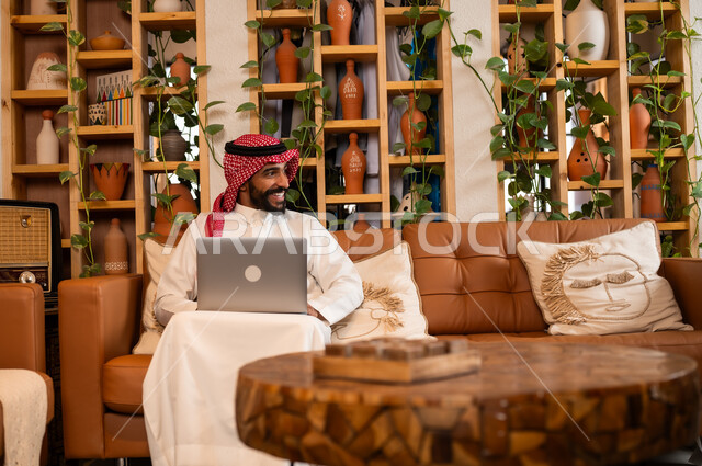  مفهوم إدارة شؤون الأعمال وتنظيمها عبر الاجهزة الحديثة، صورة مقربة لرجل عربي سعودي خليجي يرتدي الثوب التقليدي والشماغ يجلس في  محل لبيع الهدايا التذكارية التقليدية يقوم بالعمل على الحاسوب المحمول، مهن ووظائف مكتبية 