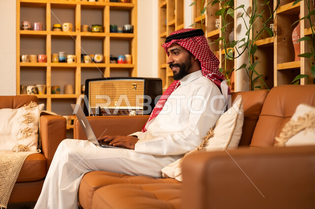  مفهوم إدارة شؤون الأعمال وتنظيمها عبر الاجهزة الحديثة، صورة مقربة من الجانب لرجل عربي سعودي خليجي يرتدي الثوب التقليدي والشماغ يجلس في  محل لبيع الهدايا التذكارية التقليدية يقوم بالعمل على الحاسوب المحمول، مهن ووظائف مكتبية 