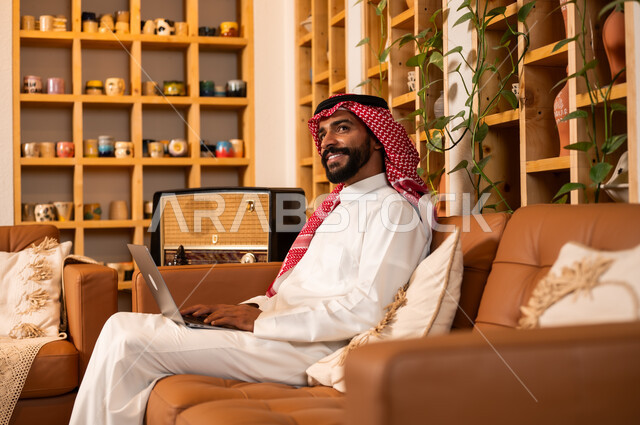  مفهوم إدارة شؤون الأعمال وتنظيمها عبر الاجهزة الحديثة، صورة مقربة من الجانب لرجل عربي سعودي خليجي يرتدي الثوب التقليدي والشماغ يجلس في  محل لبيع الهدايا التذكارية التقليدية يقوم بالعمل على الحاسوب المحمول، مهن ووظائف مكتبية 