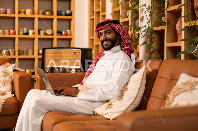  مفهوم إدارة شؤون الأعمال وتنظيمها عبر الاجهزة الحديثة، صورة مقربة من الجانب لرجل عربي سعودي خليجي يرتدي الثوب التقليدي والشماغ يجلس في  محل لبيع الهدايا التذكارية التقليدية يقوم بالعمل على الحاسوب المحمول، مهن ووظائف مكتبية 