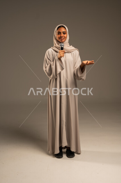 مفهوم الفن وتأثيره الايجابي على الامرأة، تقديم العروض الفنيه و الغنائيه والفقرات الترفيهية، بدء التدريب الصوتي، بورتريه لشابة عربية خليجية سعودية محجبة ترتدي العباءة الملونة تمسك بيدها المايكروفون بإيماءات الاندماج بالغناء، الجسم بالطول الكامل، خلفيه رمادية
