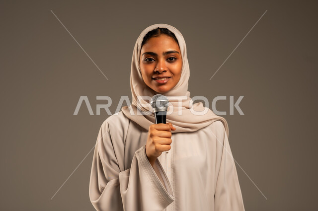 تقديم العروض الفنيه و الغنائيه والفقرات الترفيهية، مفهوم الفن وتأثيره الايجابي على الامرأة، بدء التدريب الصوتي، بورتريه مقرب لشابة عربية خليجية سعودية محجبة ترتدي العباءة الملونة تمسك بيدها المايكروفون بإيماءات الاندماج بالغناء، خلفيه رمادية