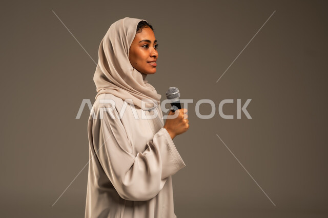تقديم العروض الفنيه و الغنائيه والفقرات الترفيهية، مفهوم الفن وتأثيره الايجابي على الامرأة، بدء التدريب الصوتي، بورتريه مقرب لشابة عربية خليجية سعودية محجبة ترتدي العباءة الملونة تمسك بيدها المايكروفون بإيماءات الاندماج بالغناء، خلفيه رمادية