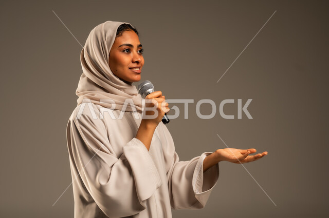 تقديم العروض الفنيه و الغنائيه والفقرات الترفيهية، مفهوم الفن وتأثيره الايجابي على الامرأة، بدء التدريب الصوتي، بورتريه مقرب لشابة عربية خليجية سعودية محجبة ترتدي العباءة الملونة تمسك بيدها المايكروفون بإيماءات الاندماج بالغناء، خلفيه رمادية