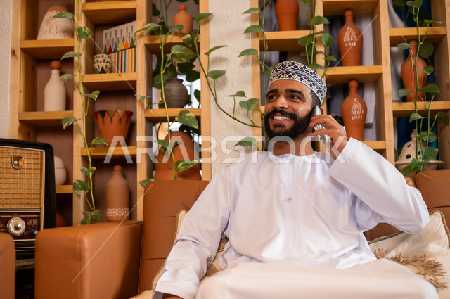 التواصل مع الأهل والأصدقاء عبر الهاتف المحمول، استخدام الأجهزة الحديثة والمتطورة، الرد على الاتصالات والمكالمات, صورة مقربة لرجل عربي خليجي عماني يرتدي الدشداشة والكمه يجلس في محل لبيع الهدايا التذكارية التقليدية يقوم بإجراء مكالمة هاتفية عبر الجوال