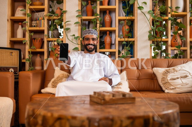 التواصل مع الأهل والأصدقاء عبر الهاتف المحمول، استخدام الأجهزة الحديثة والمتطورة، الرد على الاتصالات والمكالمات, صورة مقربة لرجل عربي خليجي عماني يرتدي الدشداشة والكمه يجلس في محل لبيع الهدايا والتحف يقوم بعرض شاشة سوداء عبر الجوال