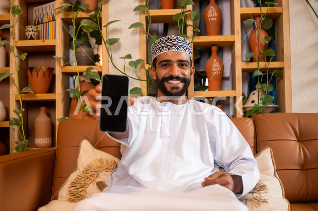التواصل مع الأهل والأصدقاء عبر الهاتف المحمول، استخدام الأجهزة الحديثة والمتطورة، الرد على الاتصالات والمكالمات, صورة مقربة لرجل عربي خليجي عماني يرتدي الدشداشة والكمه يجلس في محل لبيع الهدايا والتحف يقوم بعرض شاشة سوداء عبر الجوال