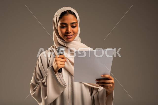 القراءة من ورقة بيضاء، تقديم العروض الفنيه و الغنائيه والفقرات الترفيهية، بدء التدريب الصوتي، بورتريه مقرب لشابة عربية خليجية سعودية محجبة ترتدي العباءة الملونة تمسك بيدها المايكروفون بإيماءات الاندماج بالغناء، خلفيه رمادية