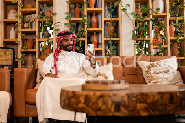 Communicating with family and friends via mobile phone, using modern and advanced devices, answering communications and calls, a close-up photo of a Saudi Gulf Arab man wearing a traditional dress sitting in a gift and antique shop holding a mobile phone in his hand.