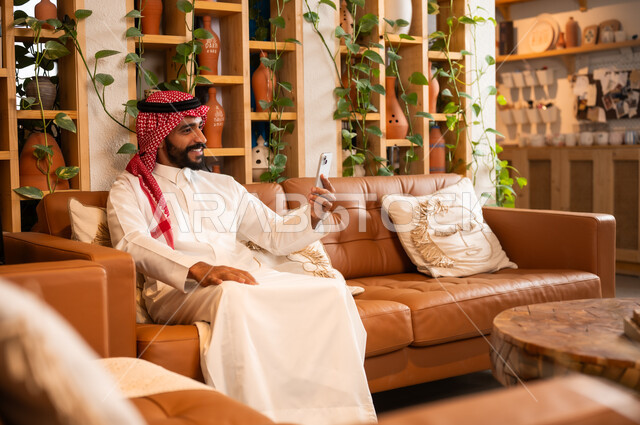 التواصل مع الأهل والأصدقاء عبر الهاتف المحمول، استخدام الأجهزة الحديثة والمتطورة، الرد على الاتصالات والمكالمات, صورة مقربة لرجل عربي خليجي سعودي يرتدي الثوب التقليدي يجلس في محل لبيع الهدايا والتحف يمسك بيده موبايل 