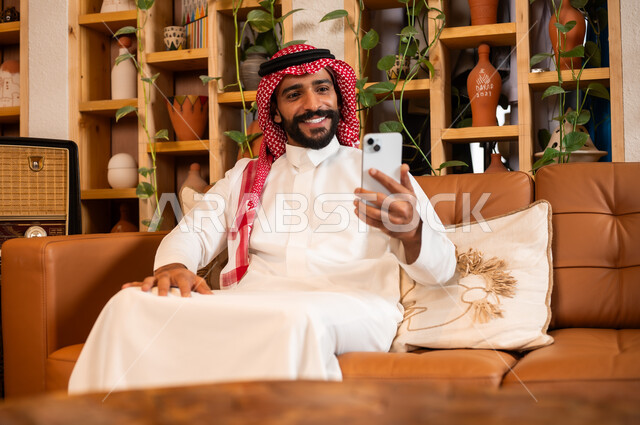 Communicating with family and friends via mobile phone, using modern and advanced devices, answering communications and calls, a close-up photo of a Saudi Gulf Arab man wearing a traditional dress sitting in a gift and antique shop holding a mobile phone in his hand.
