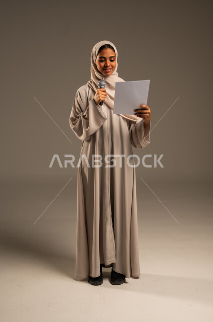 بدء التدريب الصوتي، القراءة من ورقة بيضاء، تقديم العروض الفنيه و الغنائيه والفقرات الترفيهية،  بورتريه لشابة عربية خليجية سعودية محجبة ترتدي العباءة الملونة تمسك بيدها المايكروفون بإيماءات الاندماج بالغناء، الجسم بالطول الكامل، خلفيه رمادية