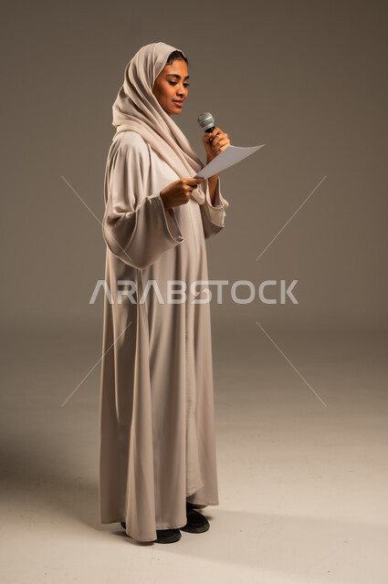 بدء التدريب الصوتي، القراءة من ورقة بيضاء، تقديم العروض الفنيه و الغنائيه والفقرات الترفيهية،  بورتريه لشابة عربية خليجية سعودية محجبة ترتدي العباءة الملونة تمسك بيدها المايكروفون بإيماءات الاندماج بالغناء، الجسم بالطول الكامل، خلفيه رمادية