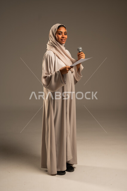 بدء التدريب الصوتي، القراءة من ورقة بيضاء، تقديم العروض الفنيه و الغنائيه والفقرات الترفيهية،  بورتريه لشابة عربية خليجية سعودية محجبة ترتدي العباءة الملونة تمسك بيدها المايكروفون بإيماءات الاندماج بالغناء، الجسم بالطول الكامل، خلفيه رمادية
