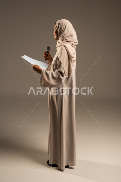 بدء التدريب الصوتي، القراءة من ورقة بيضاء، تقديم العروض الفنيه و الغنائيه والفقرات الترفيهية،  بورتريه لشابة عربية خليجية سعودية محجبة ترتدي العباءة الملونة تمسك بيدها المايكروفون بإيماءات الاندماج بالغناء، الجسم بالطول الكامل، خلفيه رمادية