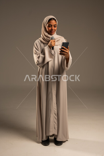 استخدام التقنيات التكنولوجية الحديثة، القراءة من الهاتف المحمول، تقديم العروض الفنيه والغنائيه والفقرات الترفيهية،  بورتريه لشابة عربية خليجية سعودية محجبة ترتدي العباءة الملونة تمسك بيدها المايكروفون بإيماءات الاندماج بالغناء، الجسم بالطول الكامل، خلفيه رمادية