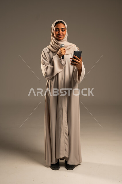استخدام التقنيات التكنولوجية الحديثة، القراءة من الهاتف المحمول، تقديم العروض الفنيه والغنائيه والفقرات الترفيهية،  بورتريه لشابة عربية خليجية سعودية محجبة ترتدي العباءة الملونة تمسك بيدها المايكروفون بإيماءات الاندماج بالغناء، الجسم بالطول الكامل، خلفيه رمادية