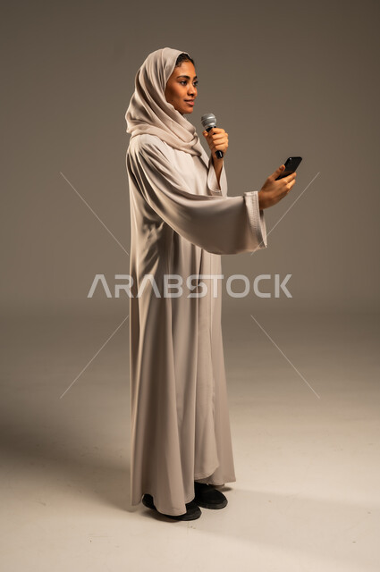 استخدام التقنيات التكنولوجية الحديثة، القراءة من الهاتف المحمول، تقديم العروض الفنيه والغنائيه والفقرات الترفيهية،  بورتريه لشابة عربية خليجية سعودية محجبة ترتدي العباءة الملونة تمسك بيدها المايكروفون بإيماءات الاندماج بالغناء، الجسم بالطول الكامل، خلفيه رمادية