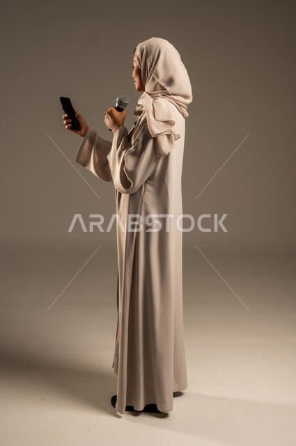 استخدام التقنيات التكنولوجية الحديثة، القراءة من الهاتف المحمول، تقديم العروض الفنيه والغنائيه والفقرات الترفيهية،  بورتريه لشابة عربية خليجية سعودية محجبة ترتدي العباءة الملونة تمسك بيدها المايكروفون بإيماءات الاندماج بالغناء، الجسم بالطول الكامل، خلفيه رمادية