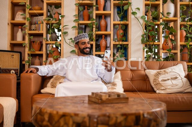 استخدام الأجهزة الحديثة والمتطورة، الرد على الاتصالات والمكالمات, صورة مقربة لرجل عربي خليجي عماني يرتدي الدشداشة والكمه يجلس في محل لبيع الهدايا والتحف يقوم بالنظر إلى الموبايل، التواصل مع الأهل والأصدقاء عبر الهاتف المحمول