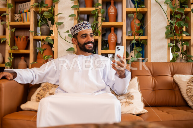 استخدام الأجهزة الحديثة والمتطورة، الرد على الاتصالات والمكالمات, صورة مقربة لرجل عربي خليجي عماني يرتدي الدشداشة والكمه يجلس في محل لبيع الهدايا والتحف يقوم بالنظر إلى الموبايل، التواصل مع الأهل والأصدقاء عبر الهاتف المحمول