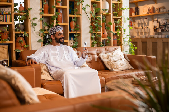 استخدام الأجهزة الحديثة والمتطورة، الرد على الاتصالات والمكالمات, صورة مقربة لرجل عربي خليجي عماني يرتدي الدشداشة والكمه يجلس في محل لبيع الهدايا والتحف يقوم بالنظر إلى الموبايل، التواصل مع الأهل والأصدقاء عبر الهاتف المحمول