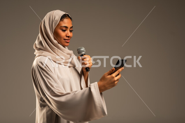 Presenting artistic and singing shows and entertainment segments, using modern technological techniques, reading from a mobile phone, close-up portrait of a veiled Saudi Arabian Gulf young woman wearing a colorful abaya holding a microphone in her hand with gestures of integration into singing, gray background