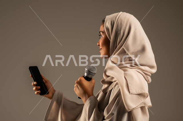 تقديم العروض الفنيه والغنائيه والفقرات الترفيهية، استخدام التقنيات التكنولوجية الحديثة، القراءة من الهاتف المحمول، بورتريه مقرب لشابة عربية خليجية سعودية محجبة ترتدي العباءة الملونة تمسك بيدها المايكروفون بإيماءات الاندماج بالغناء، خلفيه رمادية