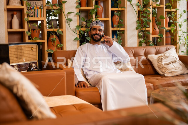 الرد على الاتصالات والمكالمات, صورة مقربة لرجل عربي خليجي عماني يرتدي الدشداشة والكمه يجلس في محل لبيع الهدايا التذكارية التقليدية يقوم بإجراء مكالمة هاتفية عبر الجوال، التواصل مع الأهل والأصدقاء عبر الهاتف المحمول، استخدام الأجهزة الحديثة والمتطورة،