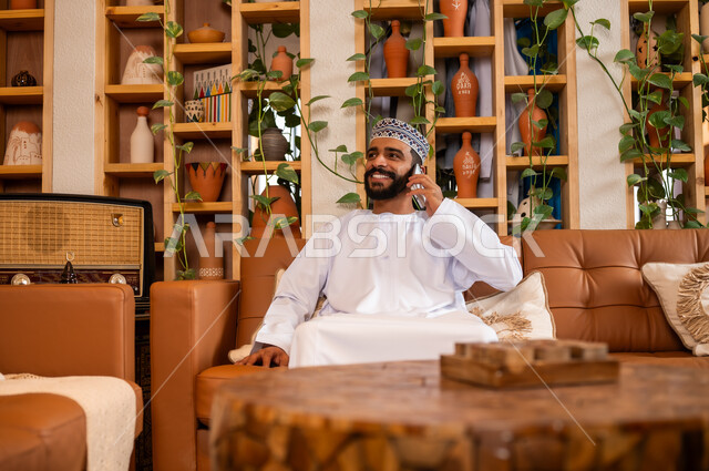 الرد على الاتصالات والمكالمات, صورة مقربة لرجل عربي خليجي عماني يرتدي الدشداشة والكمه يجلس في محل لبيع الهدايا التذكارية التقليدية يقوم بإجراء مكالمة هاتفية عبر الجوال، التواصل مع الأهل والأصدقاء عبر الهاتف المحمول، استخدام الأجهزة الحديثة والمتطورة،